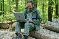 Förster sitzt auf Holzpolter mit einem Laptop in der Hand