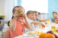 Mädchen an Essenstisch mit mehreren Kindern hält sich Gurkenscheiben vor Augen. 