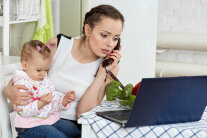 Frau sitzt mit Kleinkind an Tisch vor Notebook und hält Telefonhörer ans Ohr 
