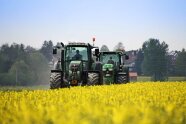 Zwei Schlepper fahren hintereinander an einem blühenden Rapsfeld entlang